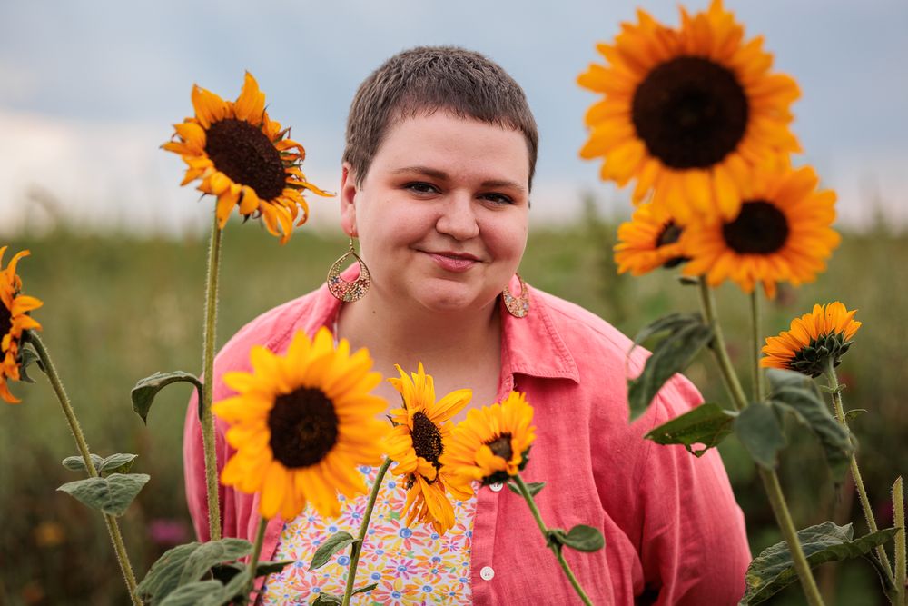 Person in coral shirt smiling among tall sunflowers in field.