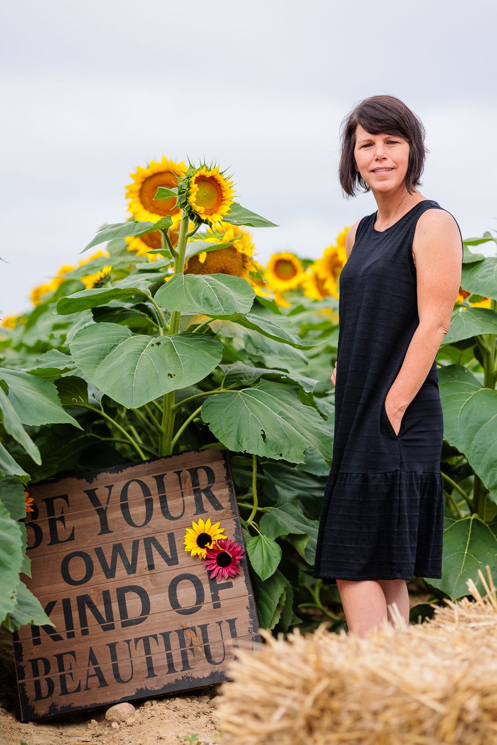 Woman in black dress standing by wooden sign in sunflower field.