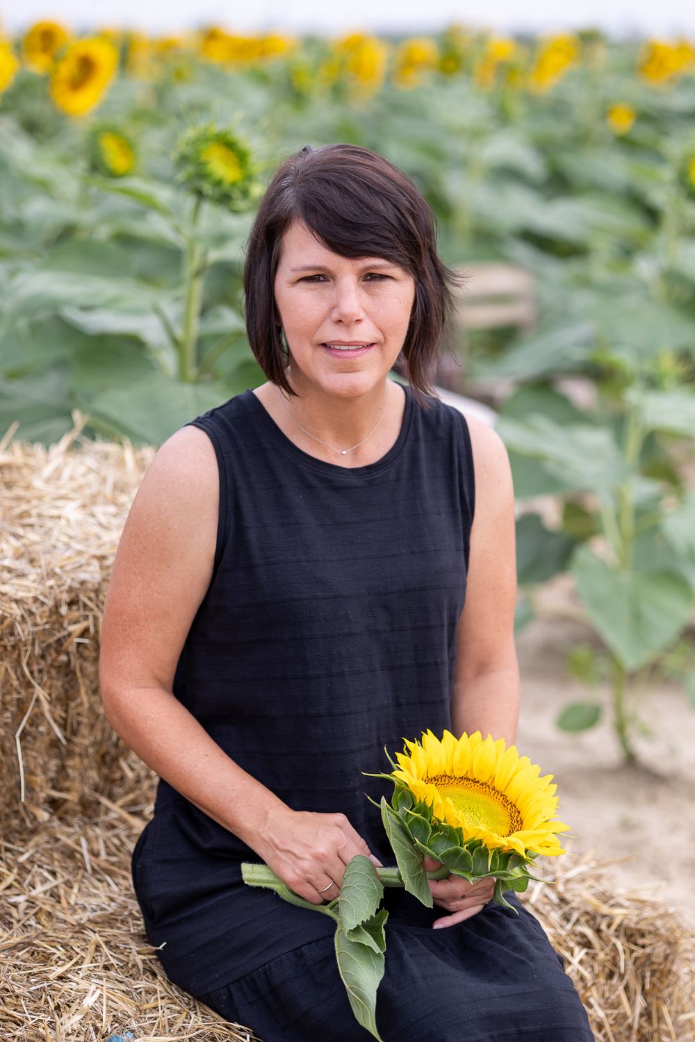 Woman in black sleeveless dress sitting on hay bale holding sunflower.