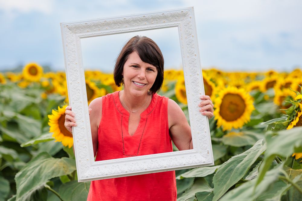 Woman in red top holding white picture frame in sunflower field.