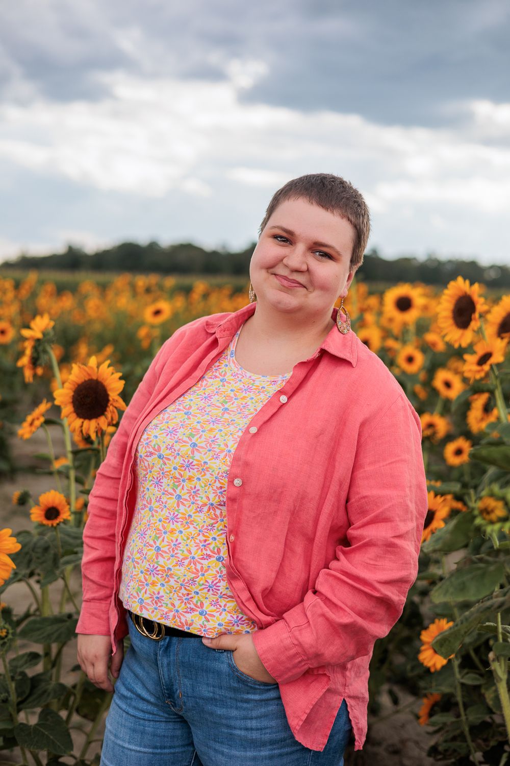 Person in pink button-up and floral top poses in blooming sunflower field.