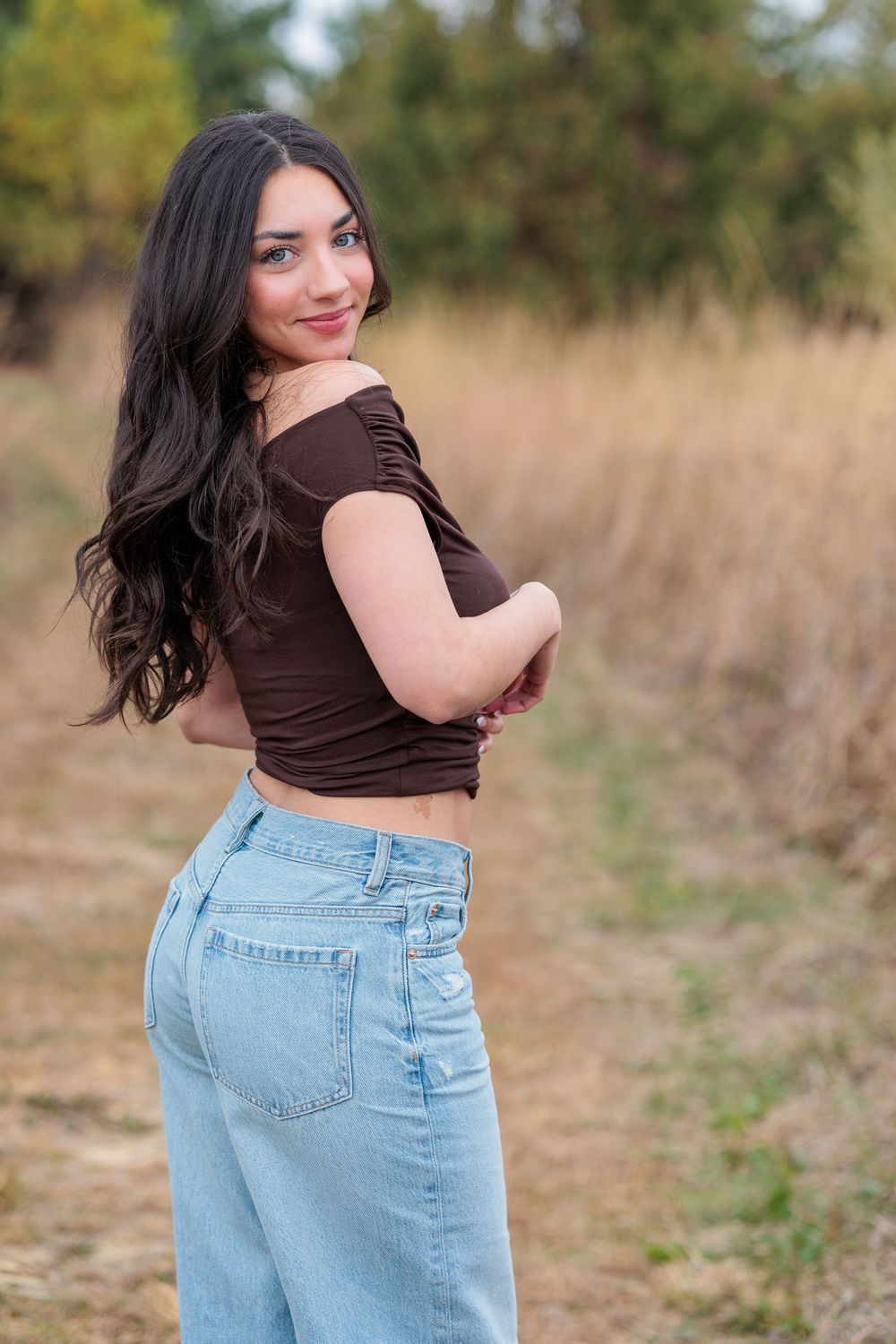 Women with beautiful long hair wearing a brown top and light wash jeans poses outdoors in a field with dried grass
