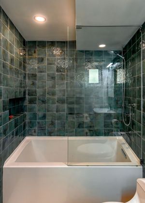 Modern bathroom featuring a deep soaking tub with glass panel, teal glazed square tiles, recessed lighting, and a handheld shower fixture.