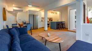 Modern open-plan living room with blue sofa, wooden coffee table, and kitchen with bar stools in a bright holiday apartment.