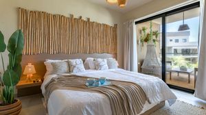 Bright bedroom with bamboo headboard, white bedding, layered textures, bedside lamp, tropical plants, and sliding doors to a sunny balcony.