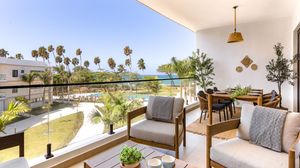 A modern apartment balcony with stylish outdoor furniture overlooks palm trees and a serene garden in warm sunlight.