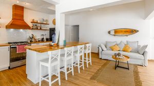 Open-plan kitchen and living room with white bar stools, wooden countertops, a surfboard wall decor, and a cozy gray sofa.
