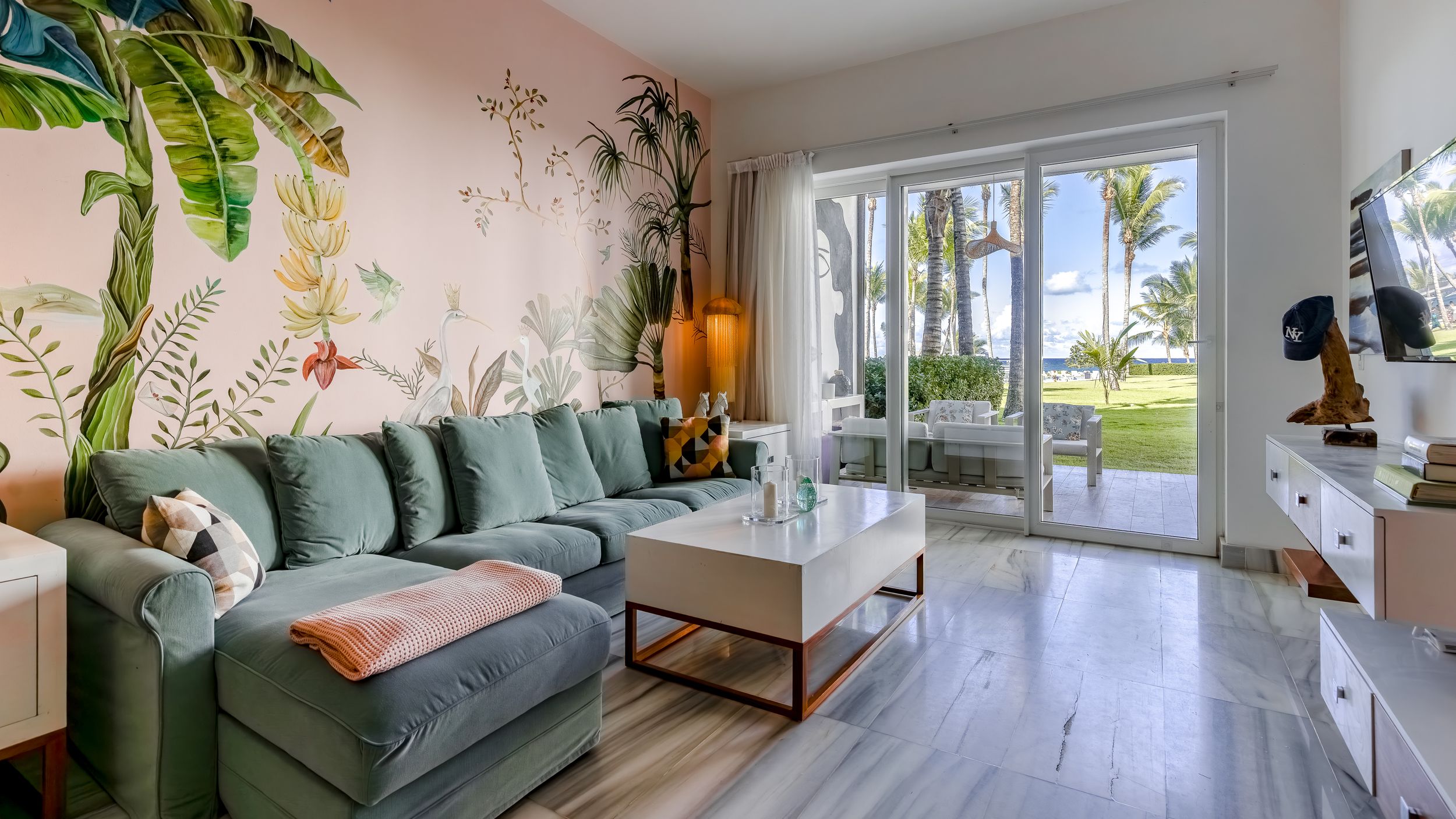 Bright living room with green sectional sofa, tropical wall mural, wood floors, and sliding doors opening to a palm tree view.