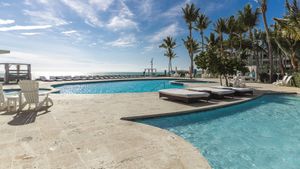 Luxury resort infinity pool with lounge chairs, palm trees, and ocean views under a partly cloudy blue sky.