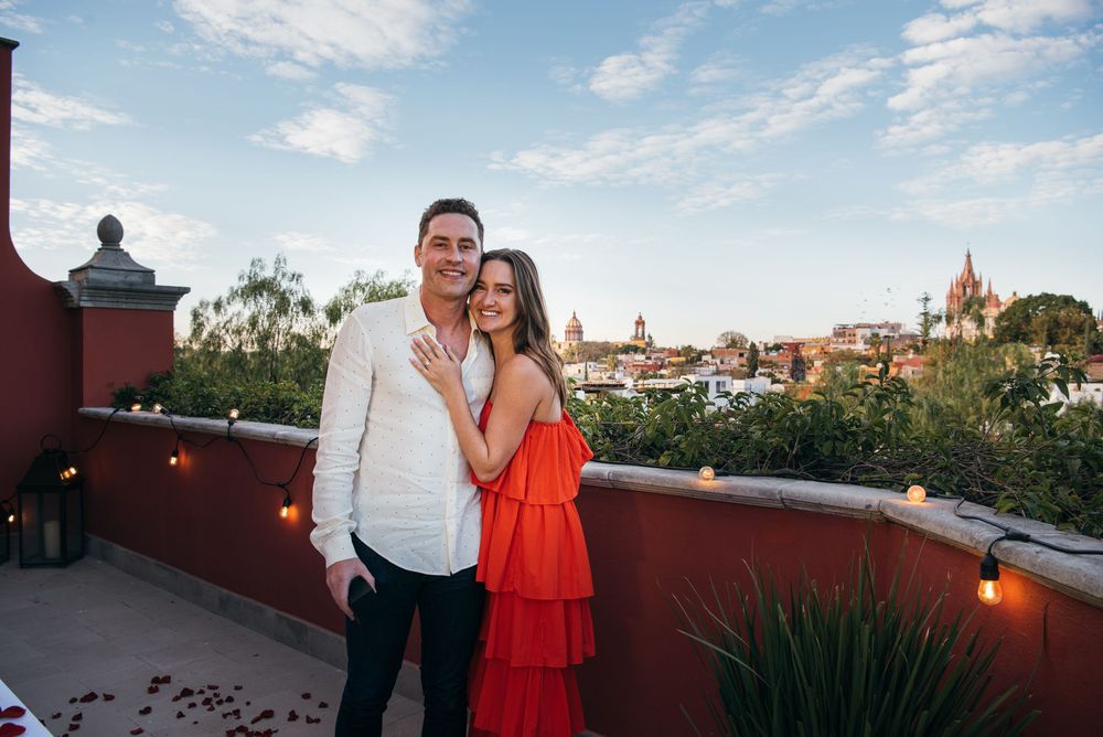 A couple stands on a rooftop terrace overlooking a scenic cityscape at sunset.
