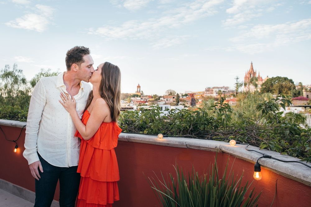 Romantic couple embraces on a rooftop terrace overlooking a historic European cityscape at sunset.