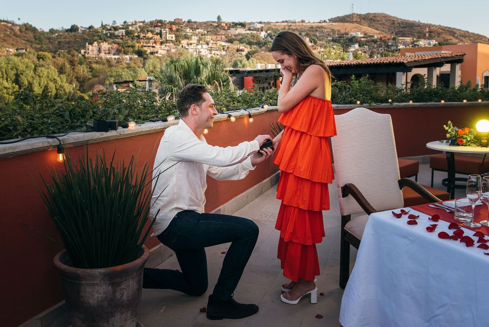 A romantic rooftop proposal scene decorated with rose petals and string lights overlooking a hillside at sunset.