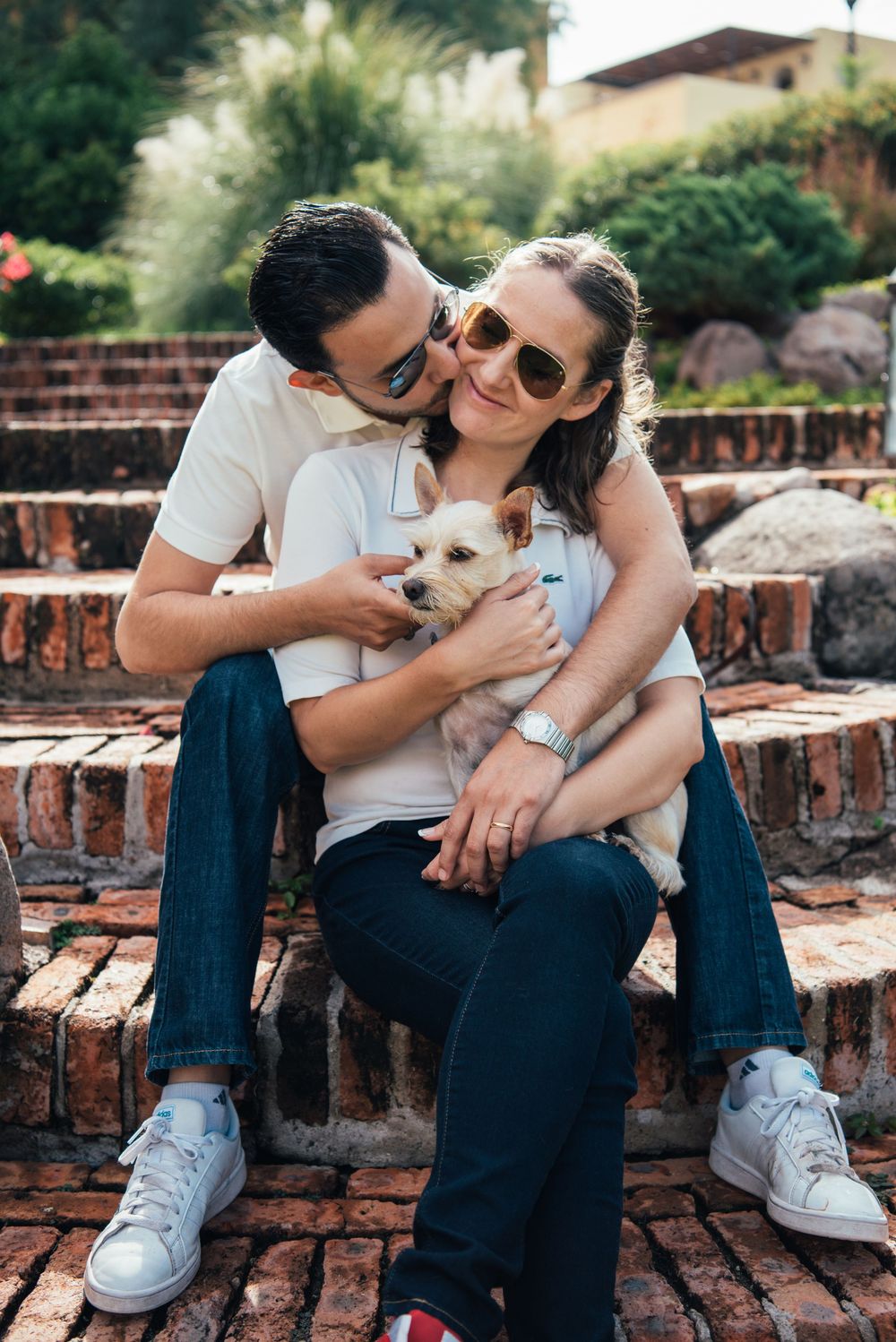 A couple sits on brick steps outdoors holding a small white dog while wearing casual clothes and sunglasses.