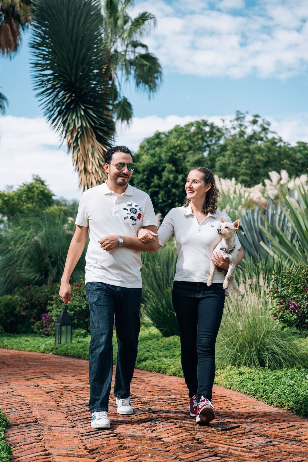 A couple in matching white shirts and jeans walks along a brick path surrounded by tropical palm trees and greenery.