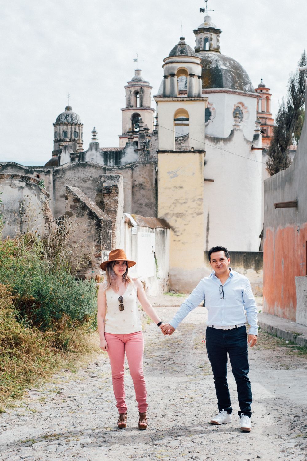 A couple walks along a rustic path with historic church domes and architecture in the background.