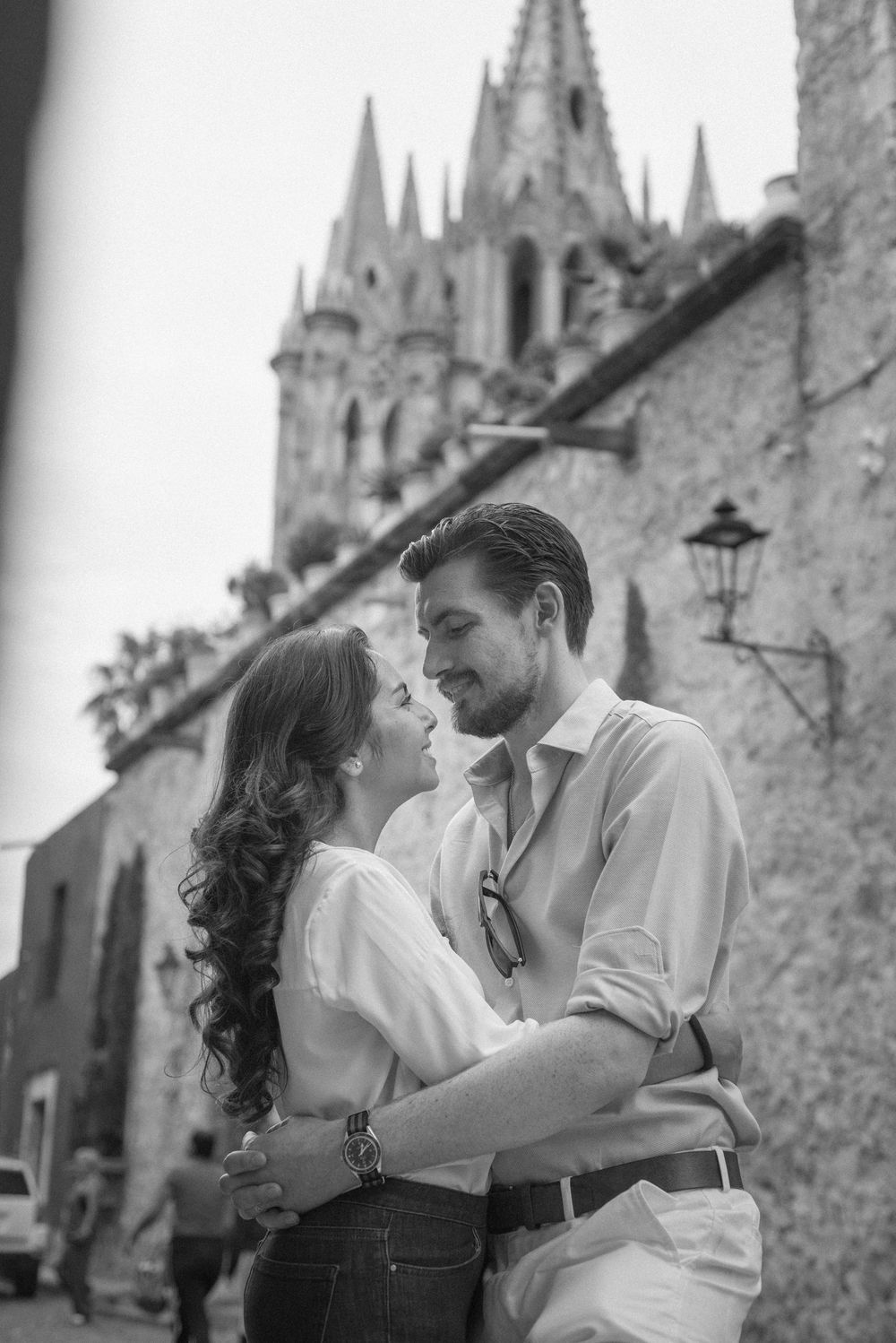 A romantic couple embraces in front of a Gothic cathedral with spires and stone architecture in black and white.