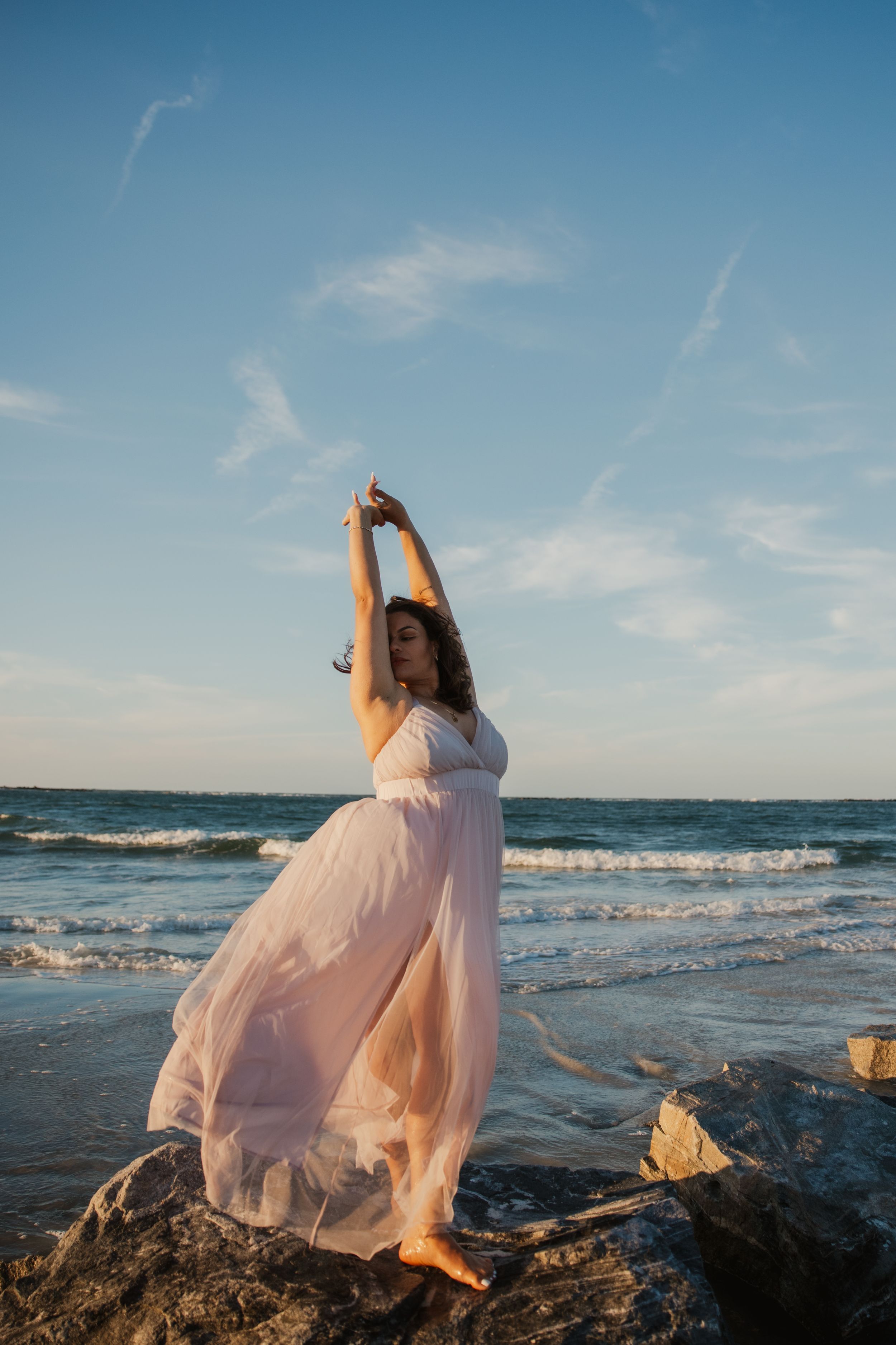 A flowing pink dress catches the ocean breeze as someone poses gracefully on coastal rocks at sunset.
