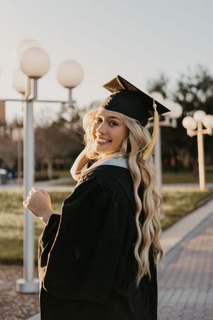 Graduate in black cap and gown poses outdoors with long wavy blonde hair against backdrop of lampposts at sunset.