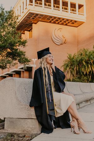 Graduate in cap and gown poses during sunset at university campus with palm trees and architectural details.