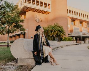A graduate in cap and gown sits on concrete steps in front of a peach-colored building with palm trees at sunset.