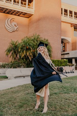 Graduate in traditional black cap and gown celebrates on campus lawn in front of peachy-colored university building.