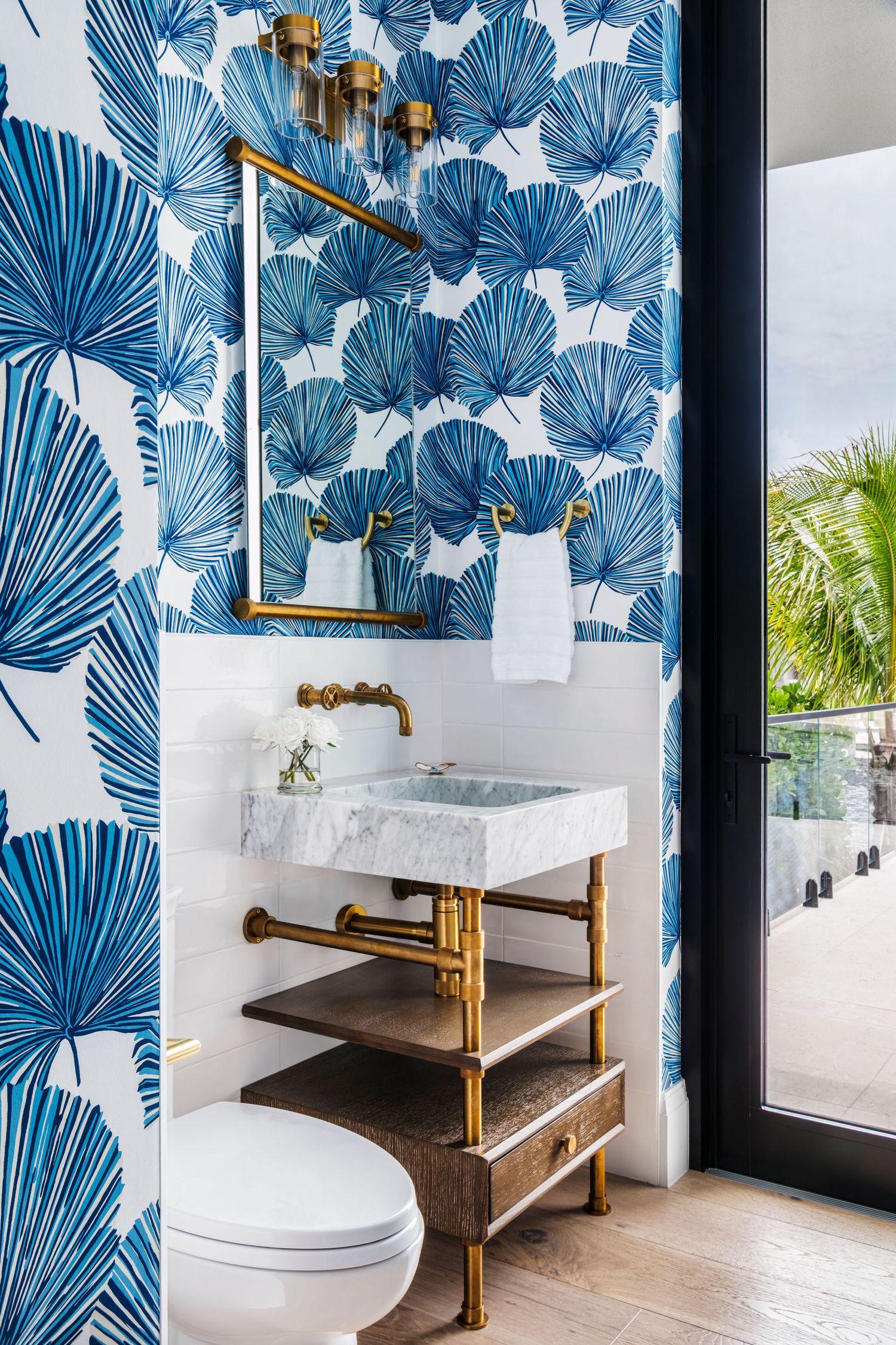 Tropical blue palm leaf wallpaper bathroom with marble sink, brass fixtures, gold-framed mirror, and white subway tile accents.