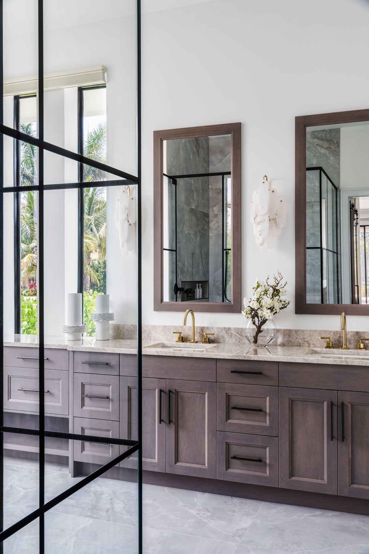 Elegant double vanity bathroom with gray cabinets, marble countertops, gold fixtures, framed mirrors, and black-framed glass shower door.