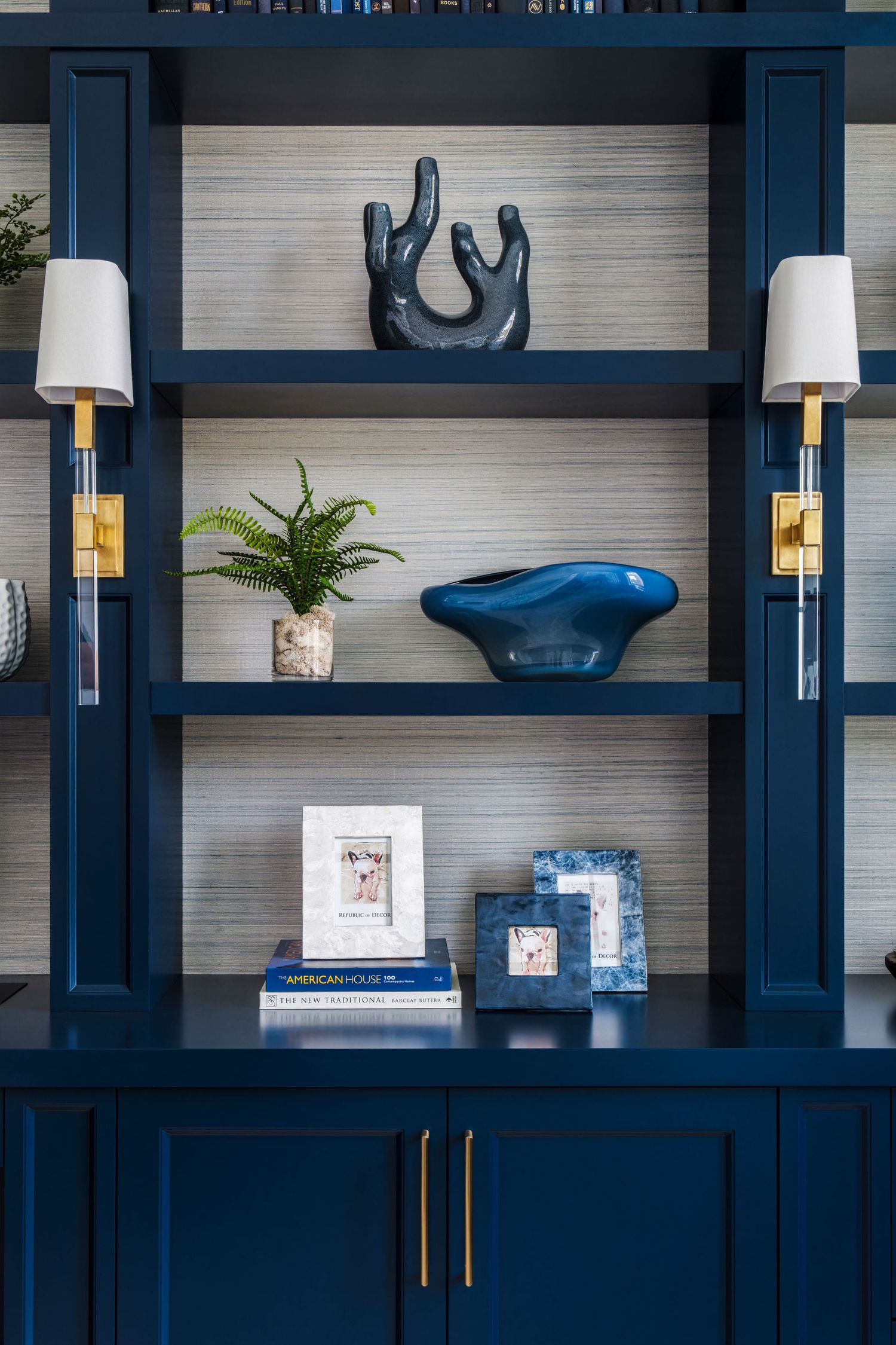 Navy blue built-in bookshelf with gold hardware, decorative objects, fern plant, and wall sconces on grasscloth wallpaper.
