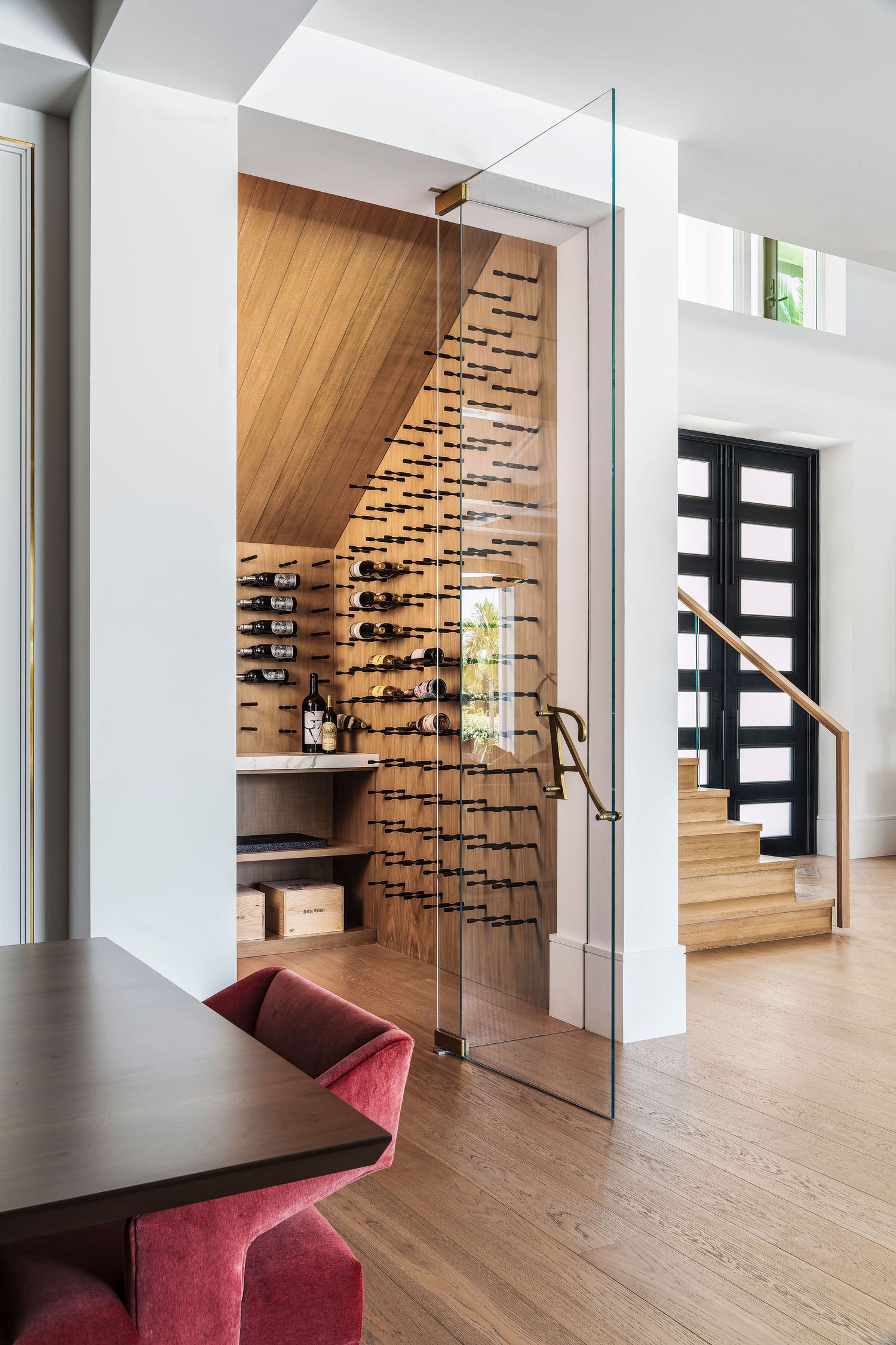Modern home wine cellar under stairs with glass door, wooden walls, and bottle racks beside a dining area.