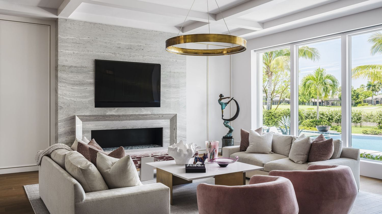 Modern living room with gray sofas, marble fireplace, gold ring chandelier, and large windows overlooking palm trees.