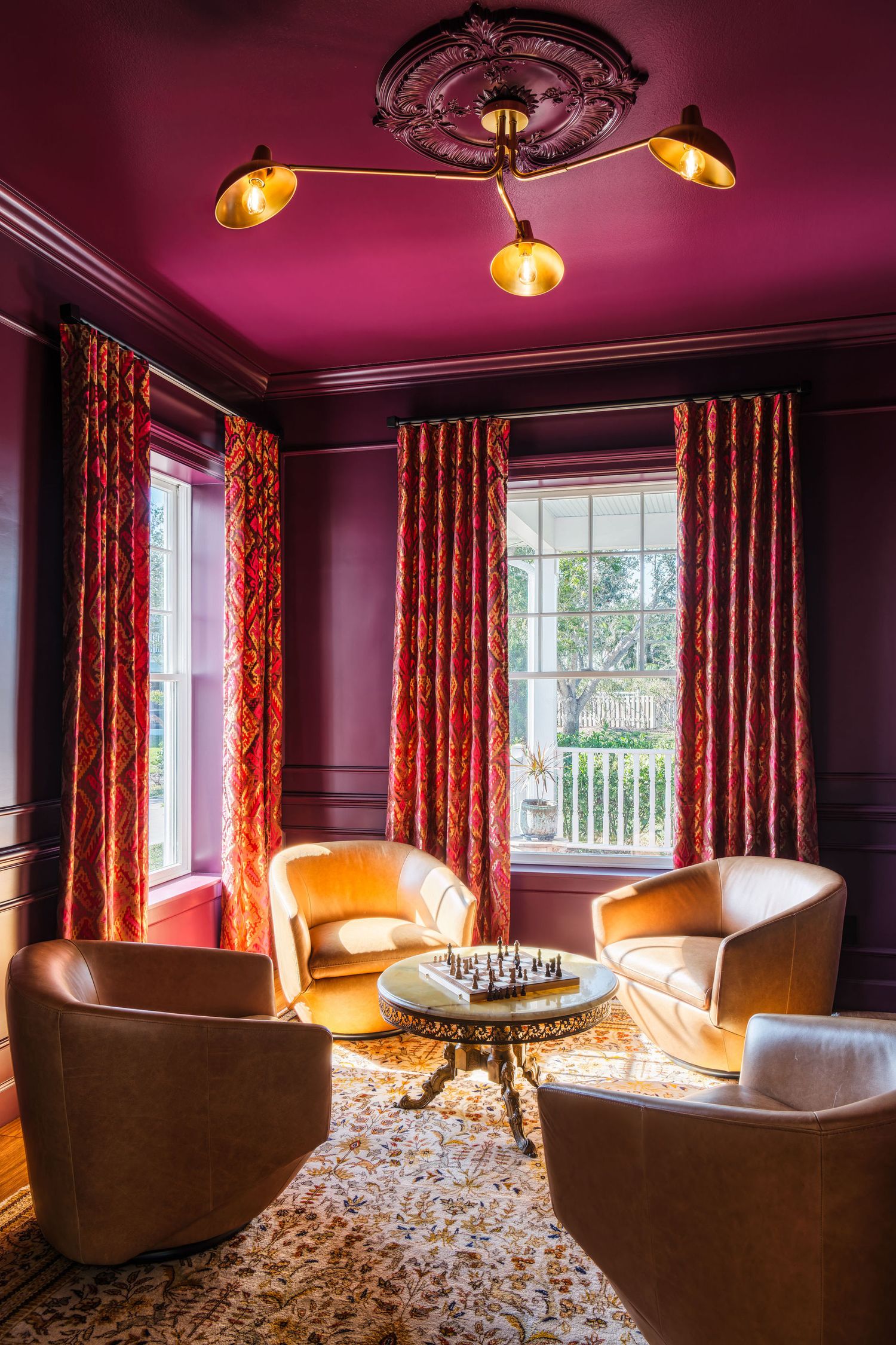 Elegant sitting room with magenta walls, gold ceiling light, tan armchairs, patterned curtains, and a chess set on a round table.