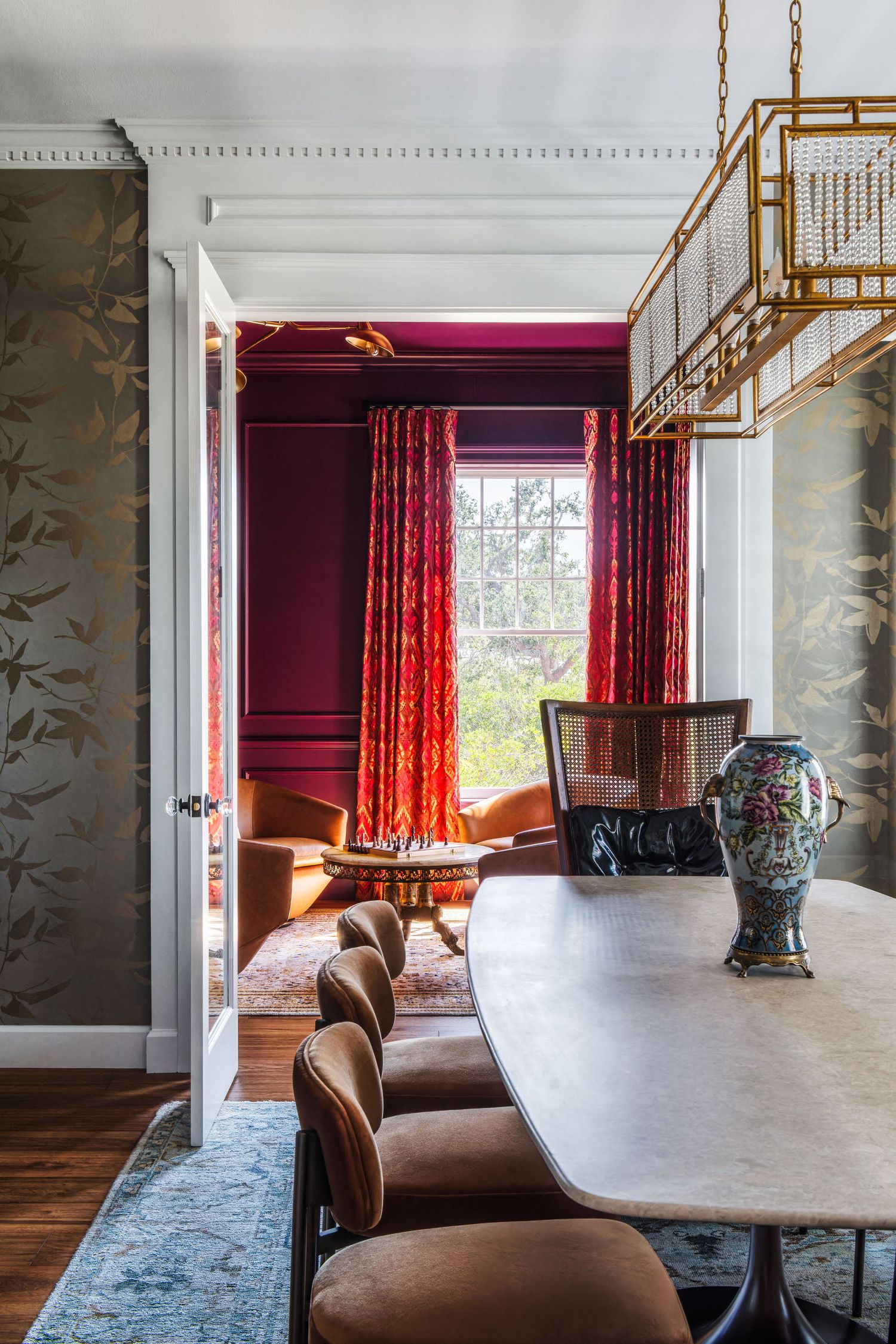 Elegant dining room with burgundy accent wall, red floral curtains, gold chandelier, marble table, and decorative vase.