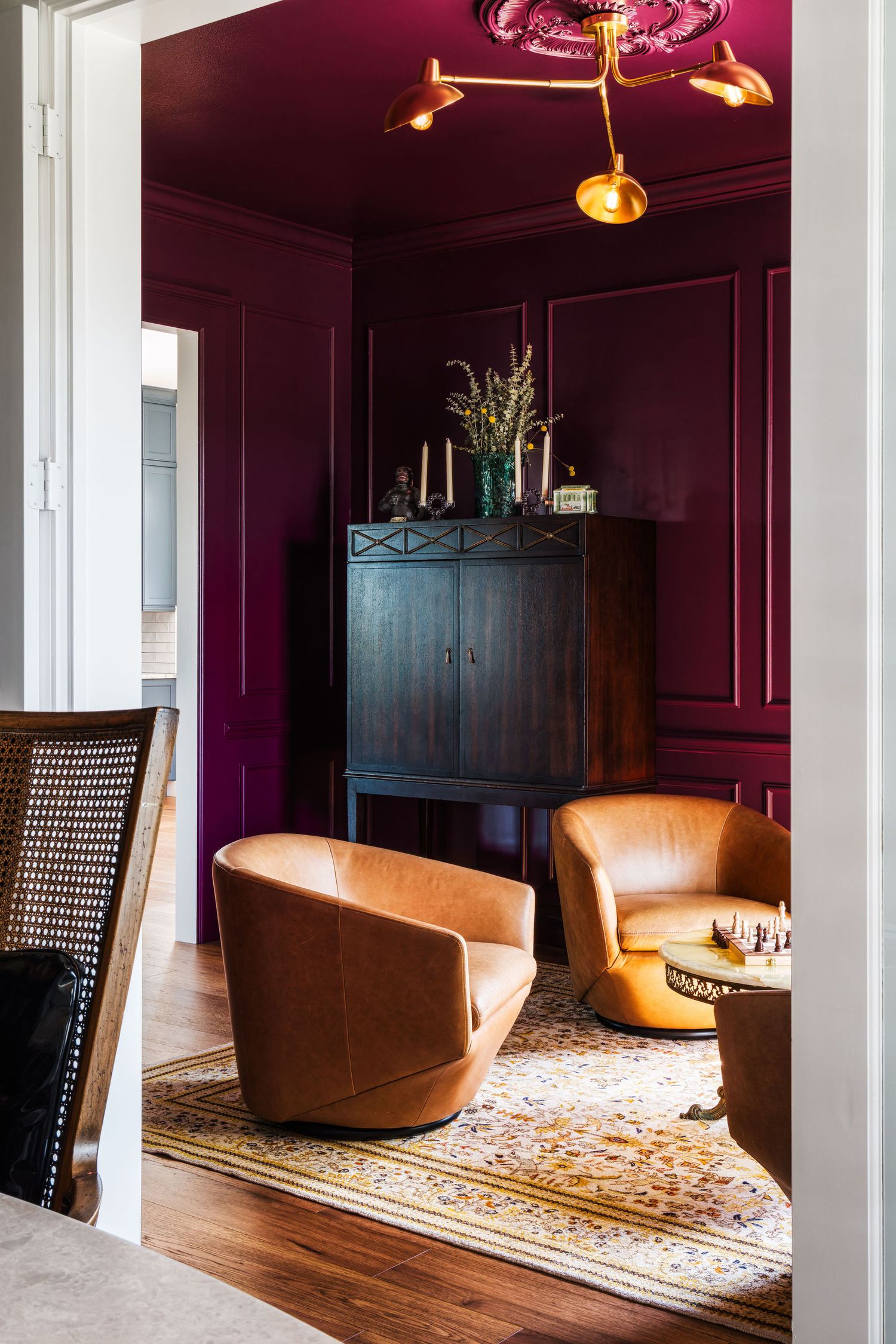 Elegant sitting room with deep burgundy walls, tan leather chairs, dark wood cabinet, and gold chandelier accents.