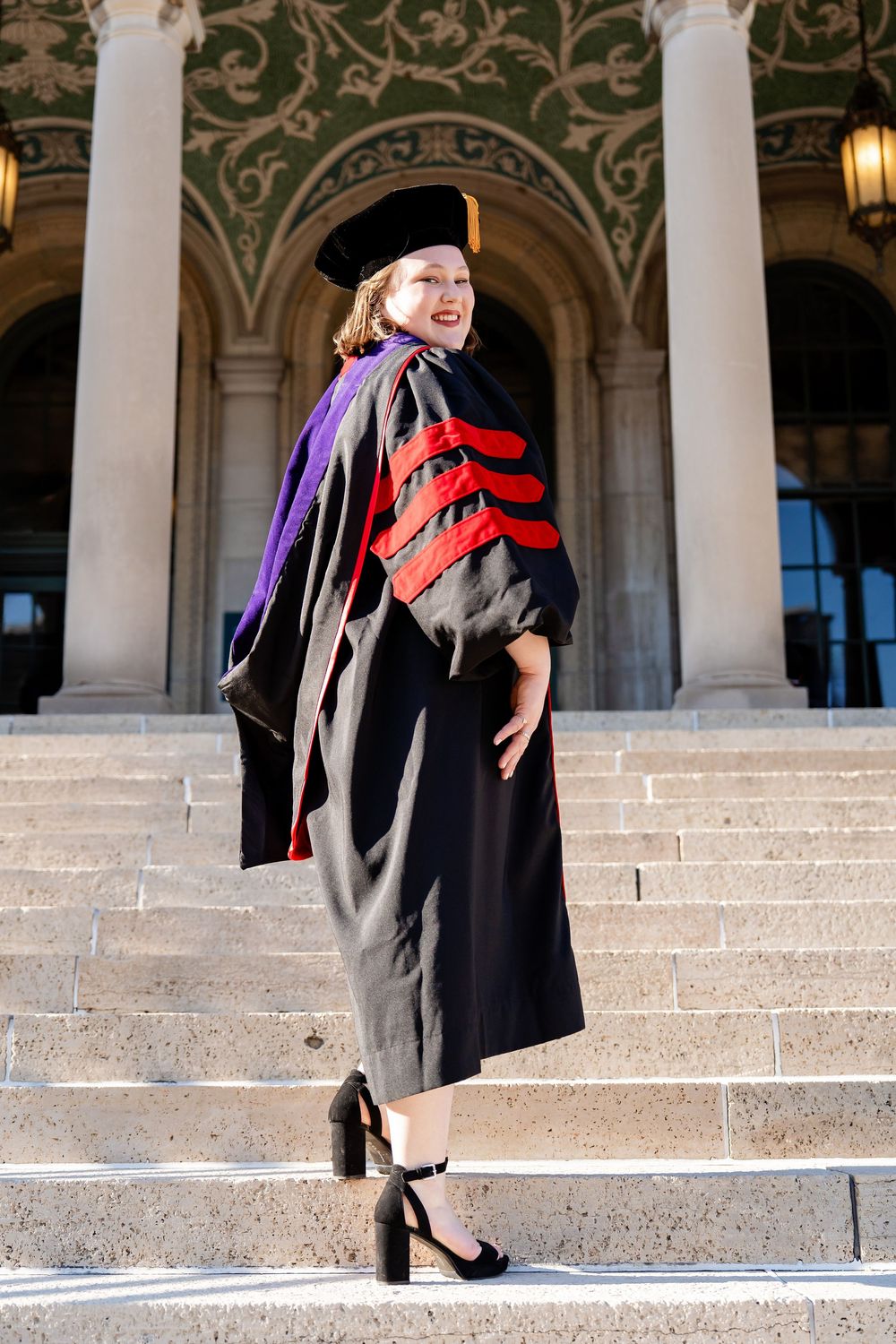 A graduate wearing full regalia on a staircase looking over her shoulder taken by Milwaukee photographer Rachel Dickman.