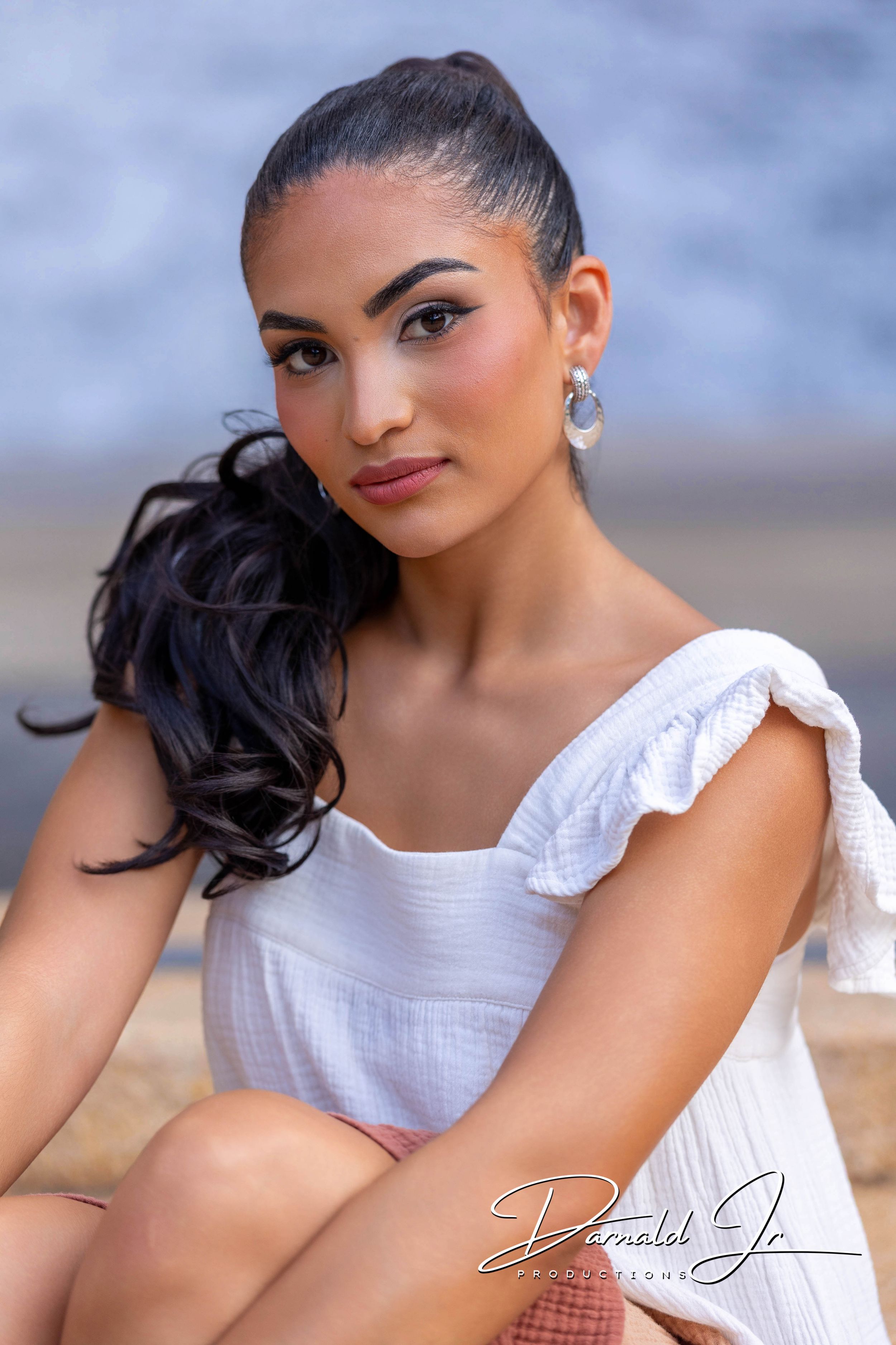 Young woman in white ruffled dress with dark curly ponytail and hoop earrings poses elegantly outdoors.