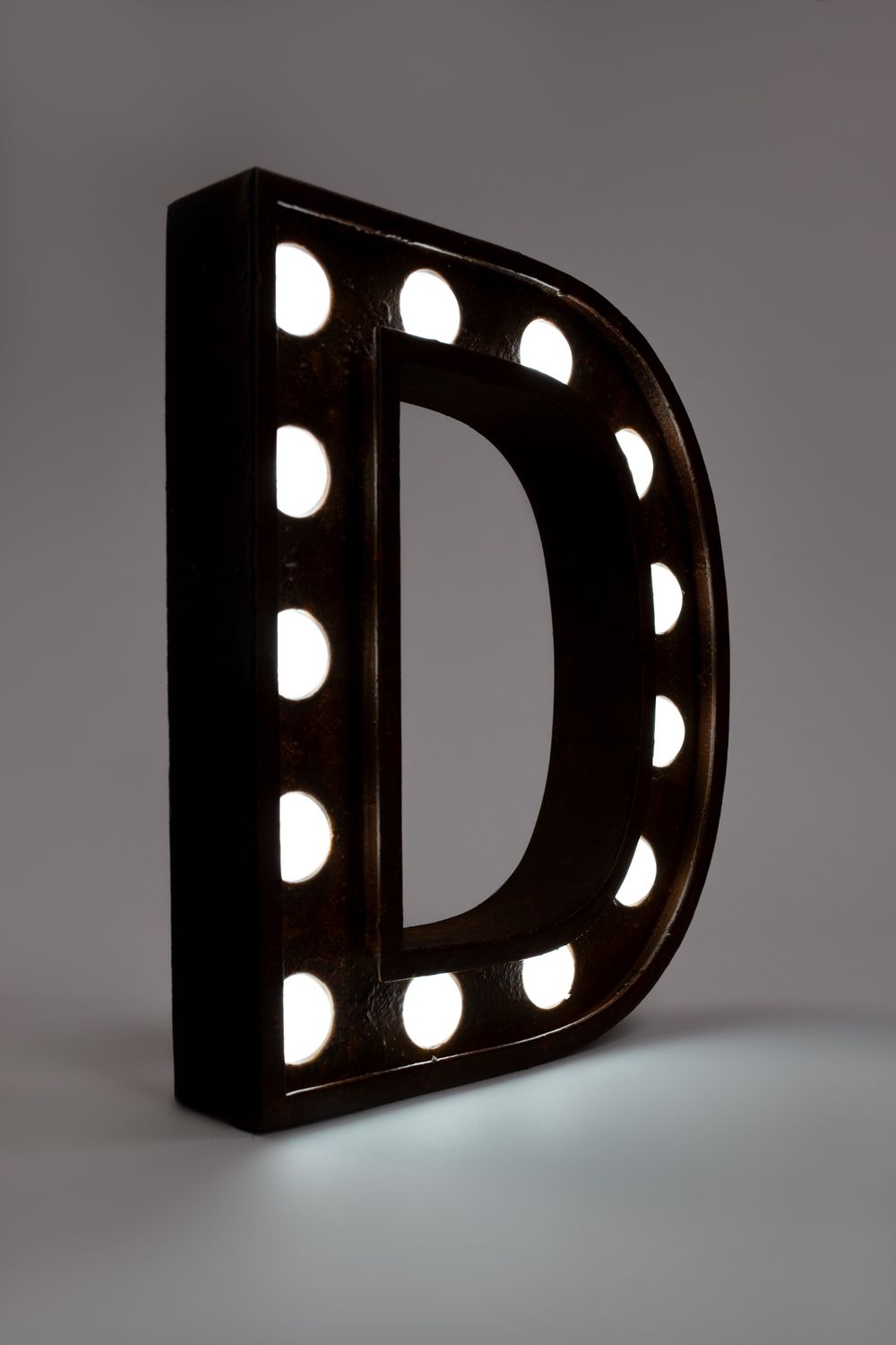 An illuminated letter D marquee sign with glowing white bulbs against a dark background stands upright in dramatic lighting.