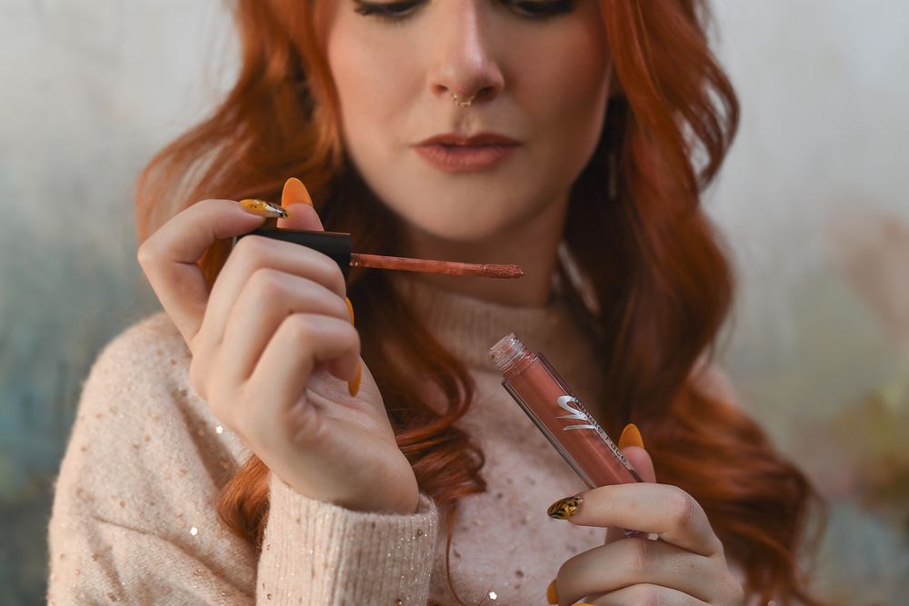 A person with long wavy auburn hair wearing a beige sweater holds art supplies in their hands.
