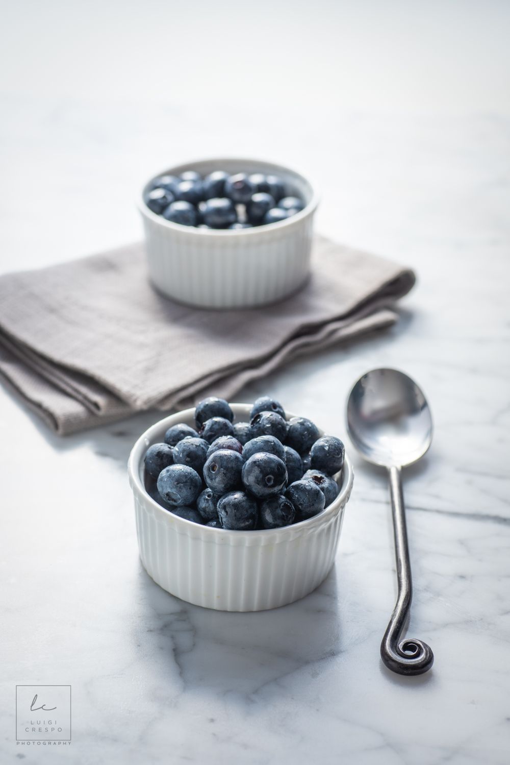 Fresh blueberries served in white ramekins on marble surface with vintage spoon and gray linen napkin.