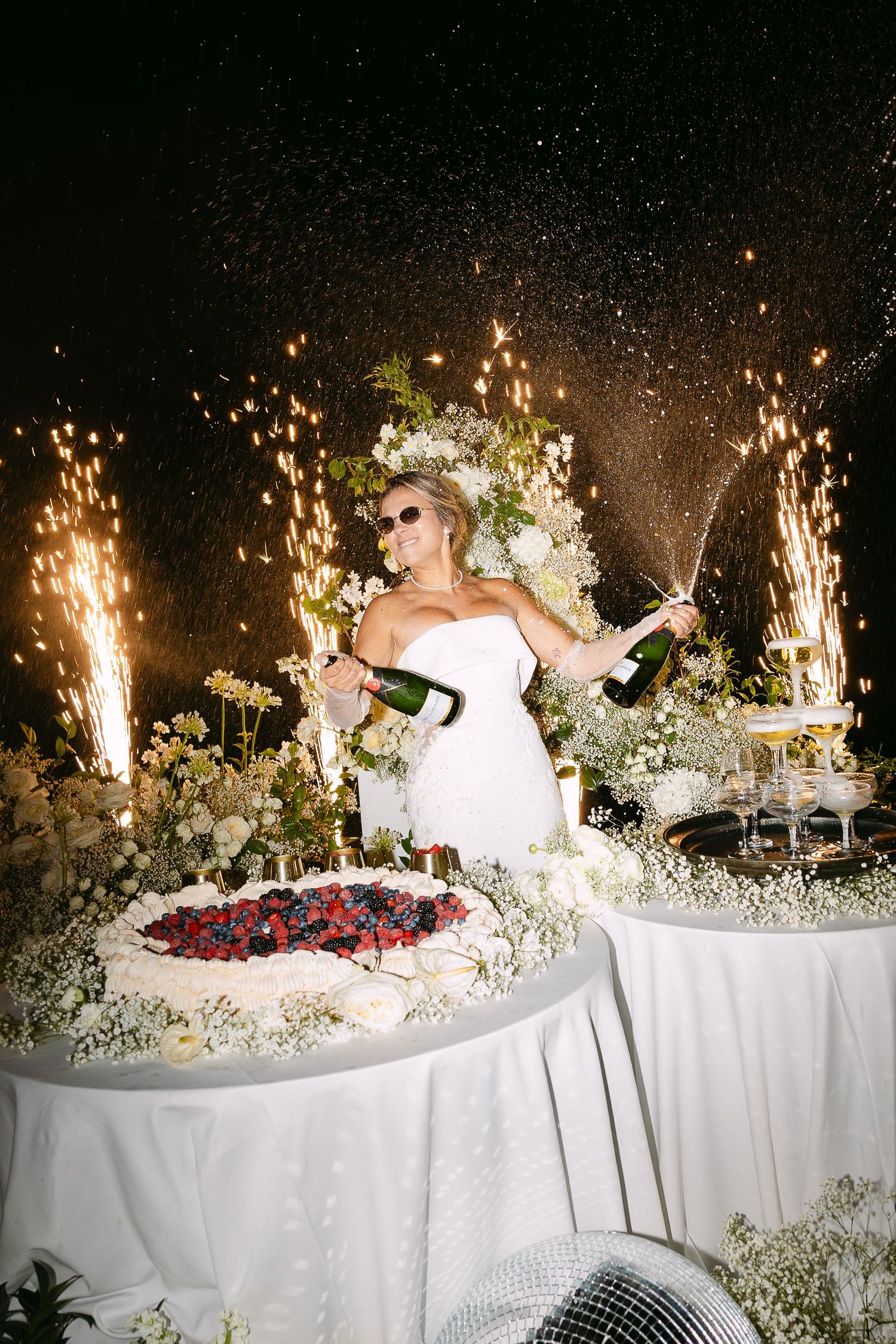Bride in white strapless gown sprays champagne near wedding cake surrounded by sparklers and white floral arrangements.