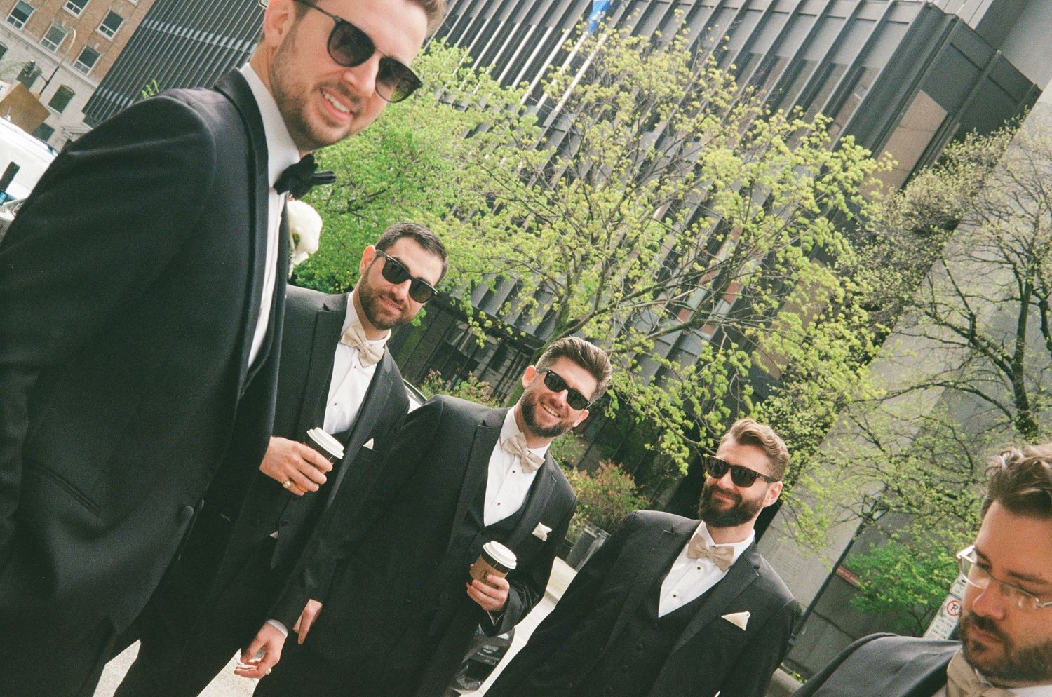 A group in black tuxedos and sunglasses stand together outdoors on a sunny day with greenery in the background.