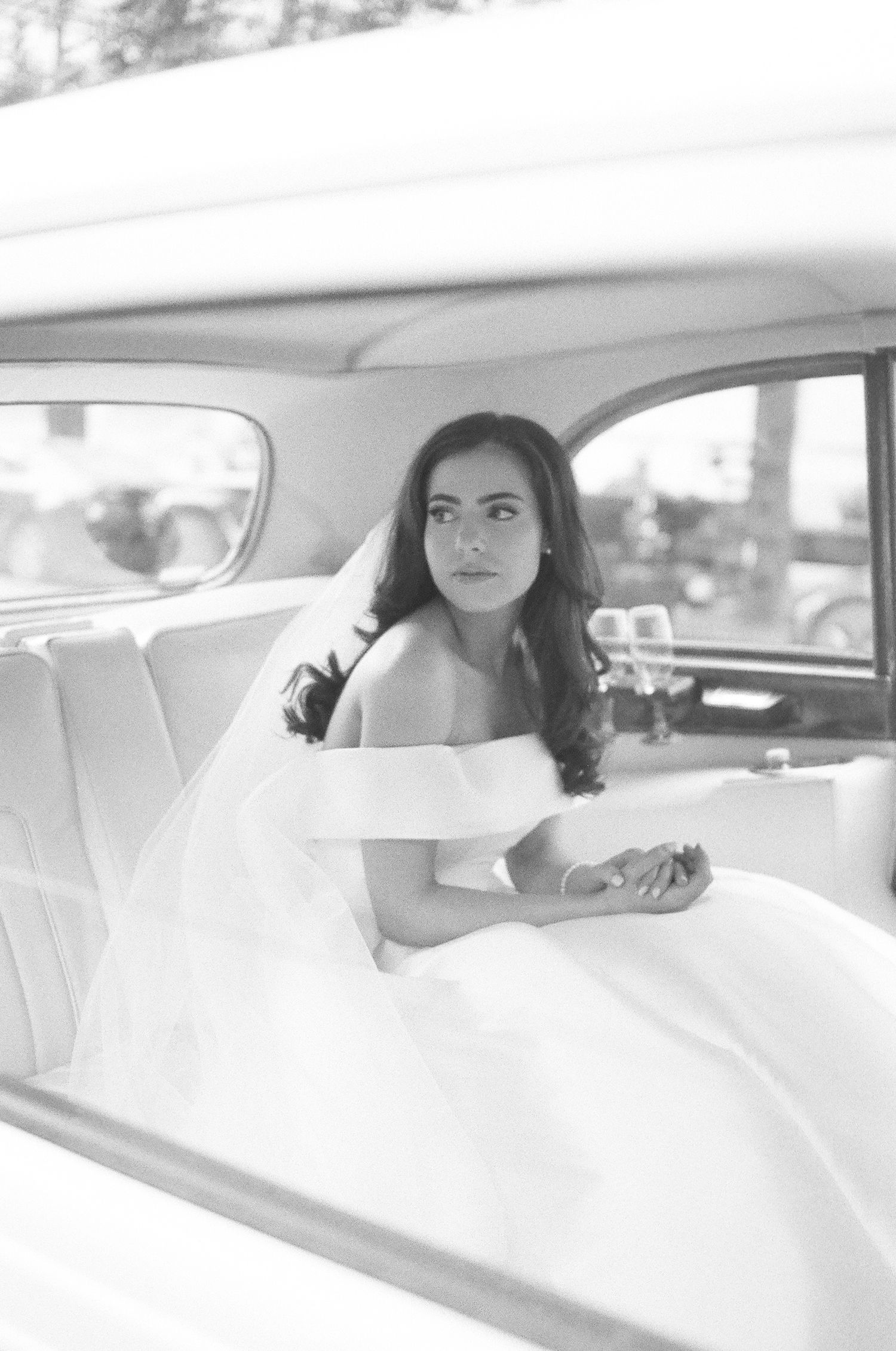 A bride in a white wedding dress sits elegantly in the backseat of a classic car looking out the window.
