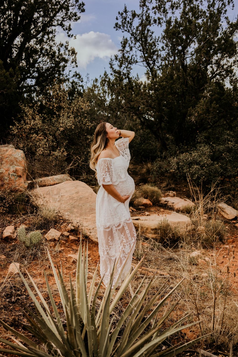 Maternity photoshoot in nature featuring white lace dress and desert landscape with agave plant.