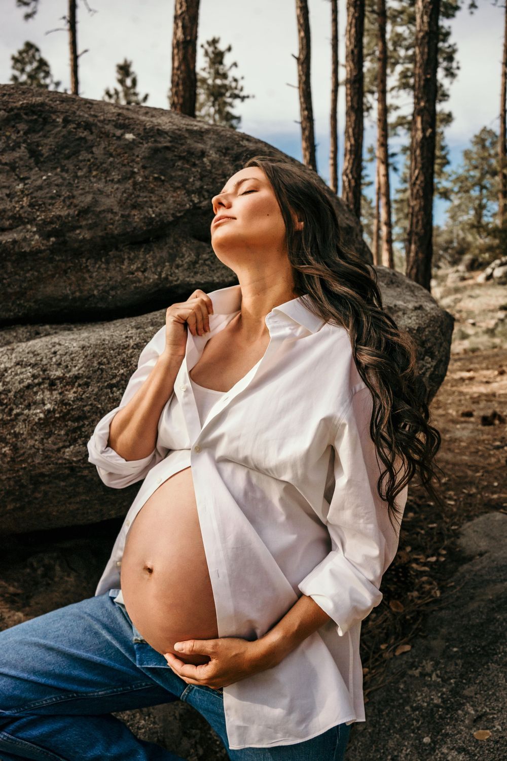 Artistic maternity photo with white button-down shirt against natural rock formations and pine trees.