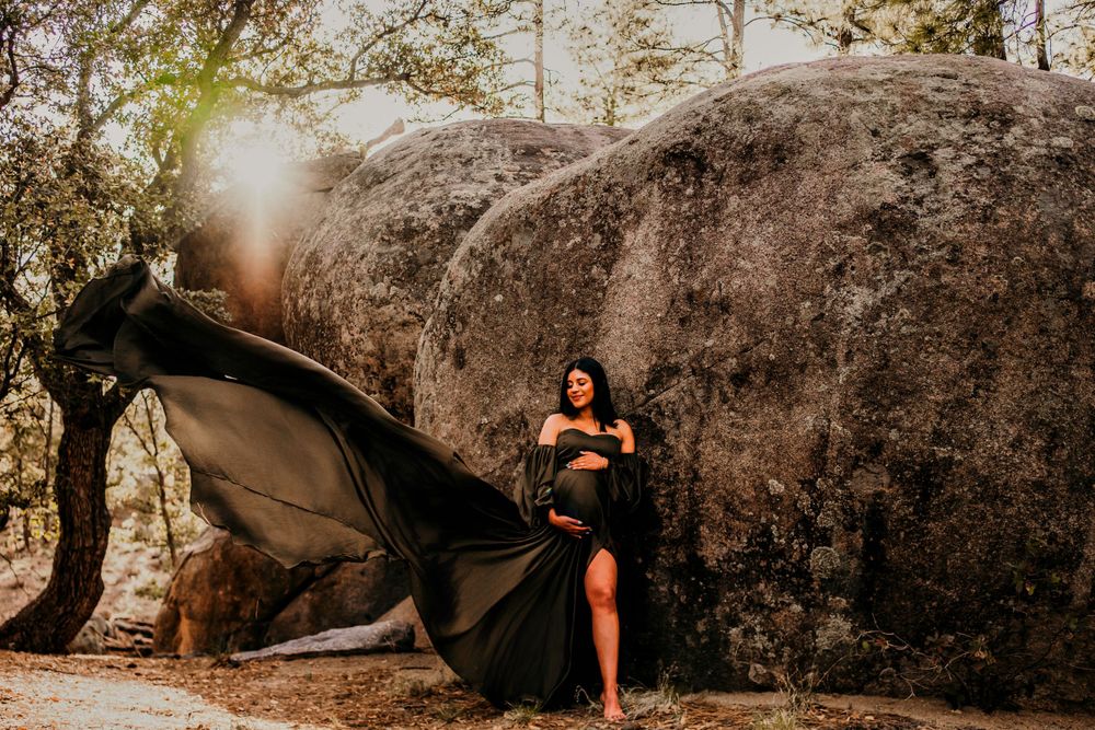 Dramatic maternity photo with flowing black dress against massive boulder in golden light.