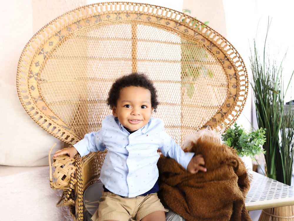 Smiling toddler boy sitting in a wicker peacock chair holding stuffed animals, wearing a light blue shirt and khaki pants.