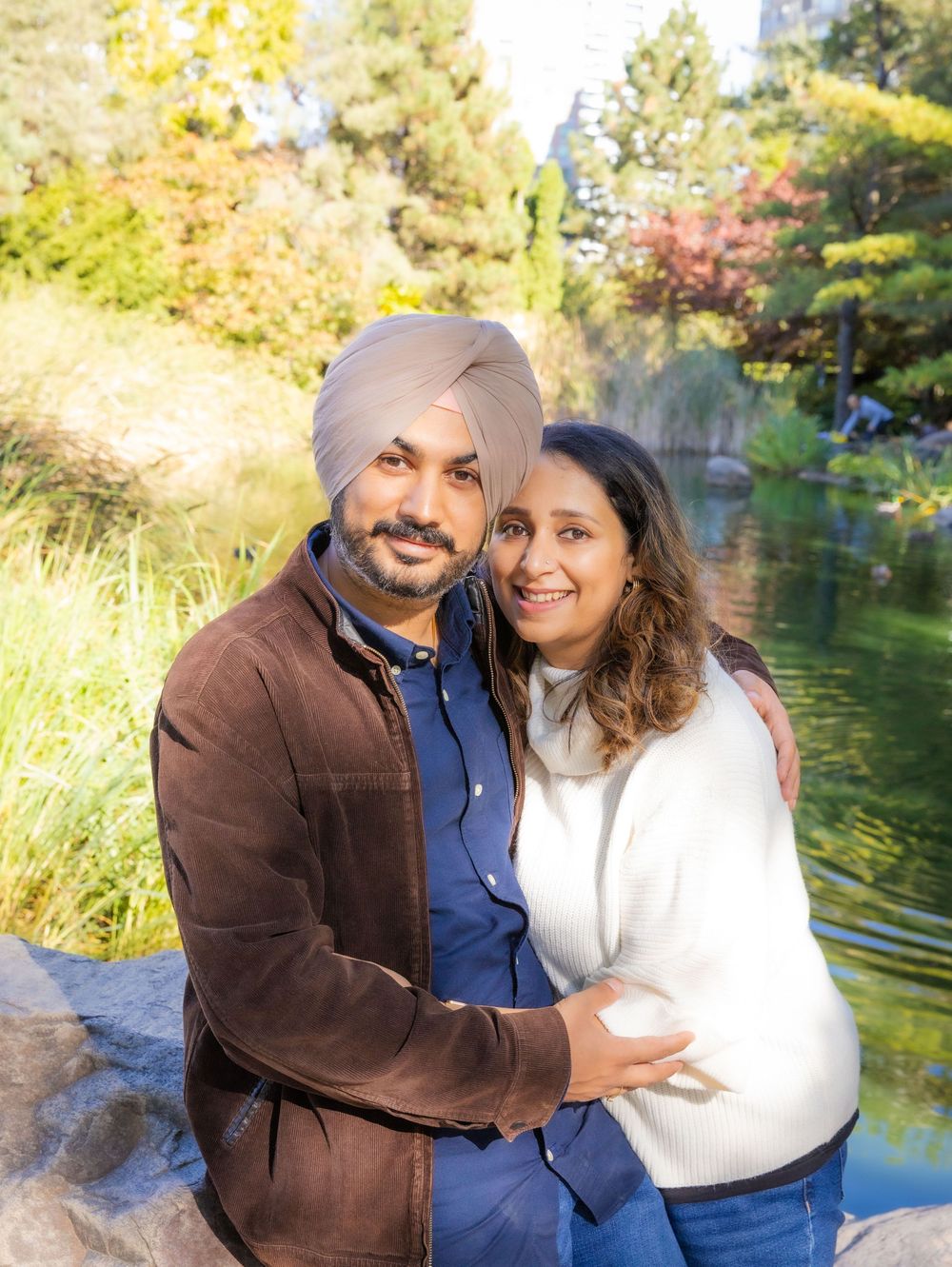 A Sikh man in a brown jacket and turban embraces a woman in a white sweater near a scenic garden pond.