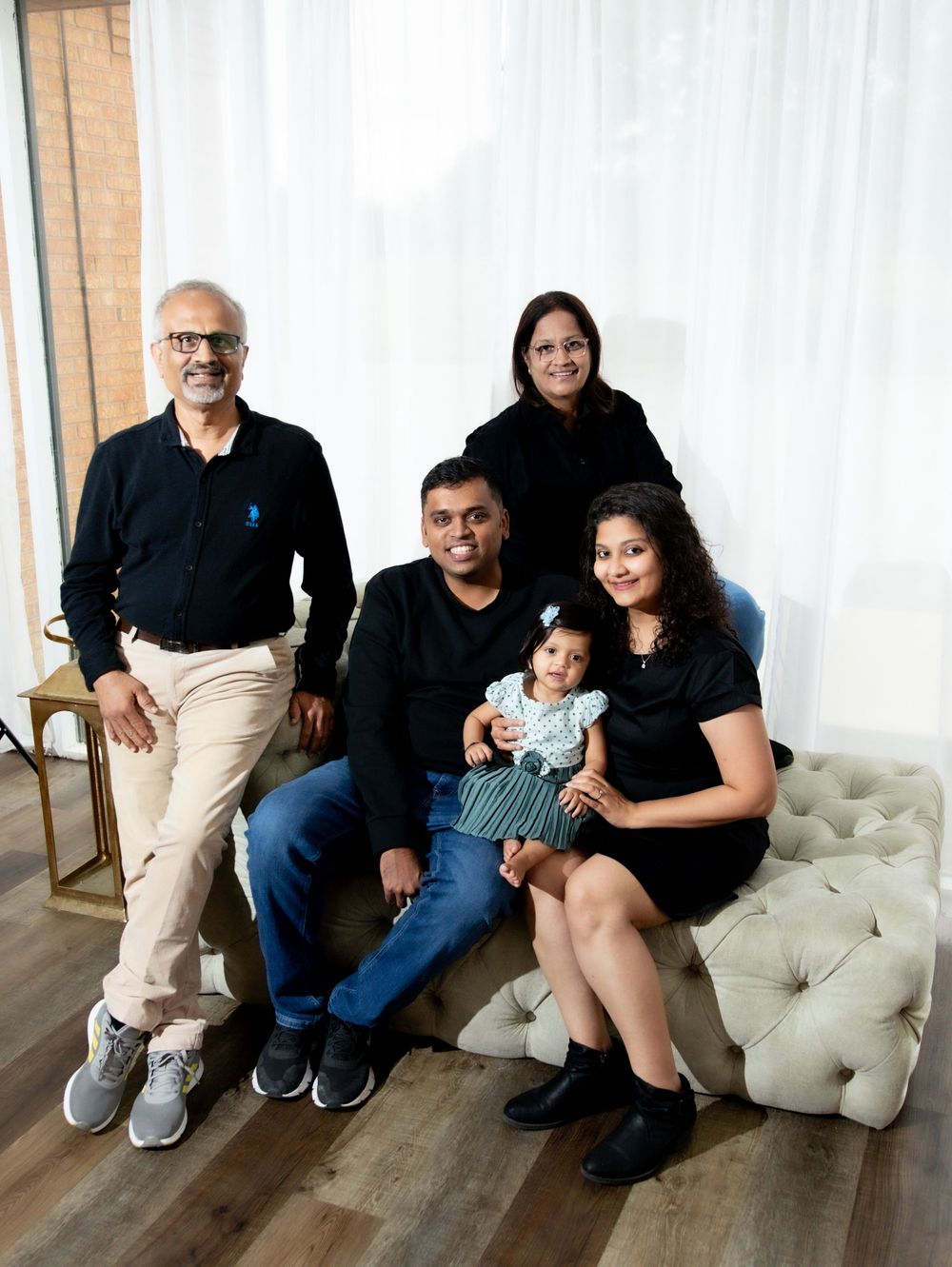 Multi-generational South Asian family portrait with five members including a baby, posing together on a tufted bench indoors.