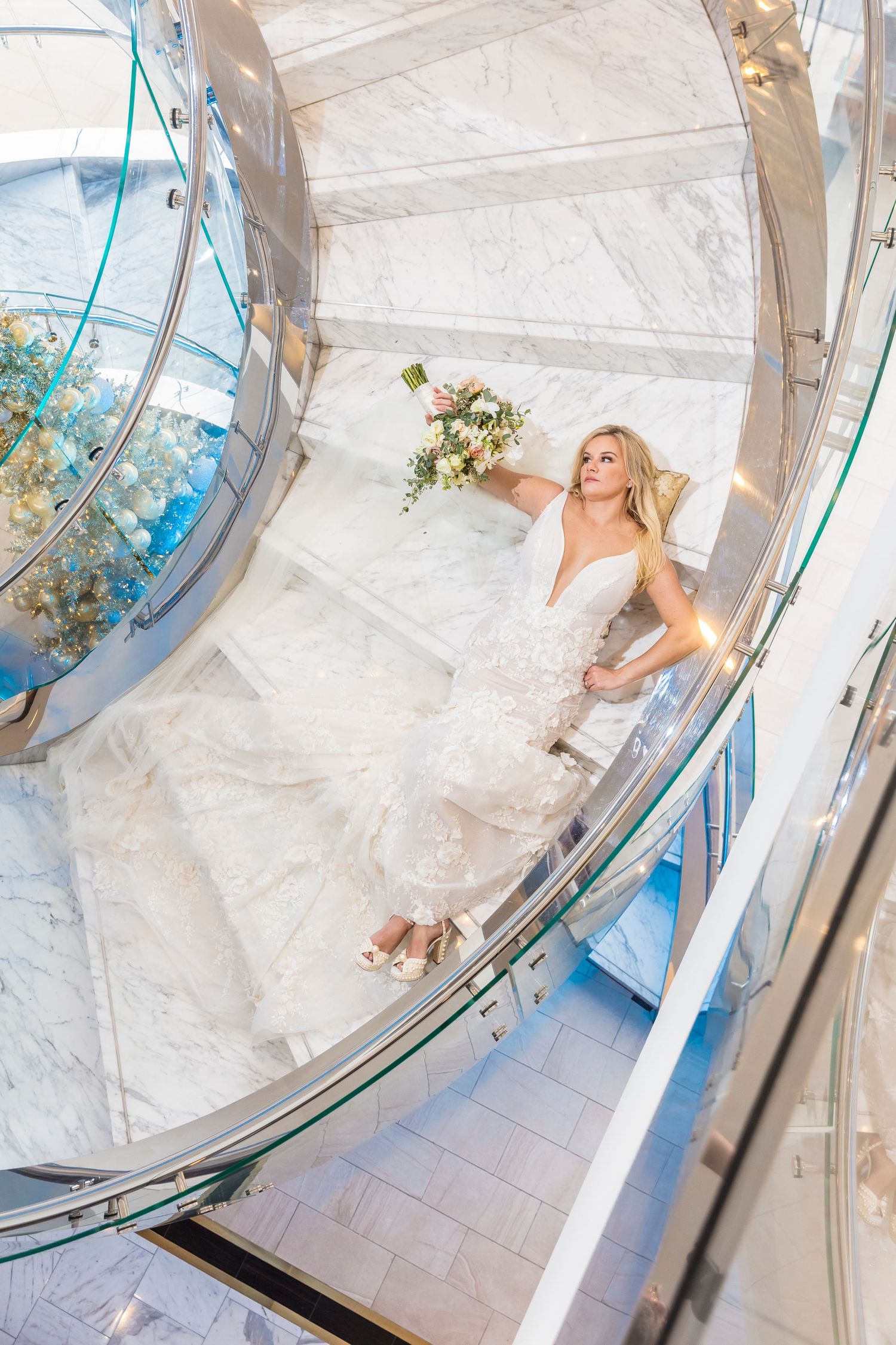 Bride in white gown holding bouquet poses on marble spiral staircase, viewed from above.