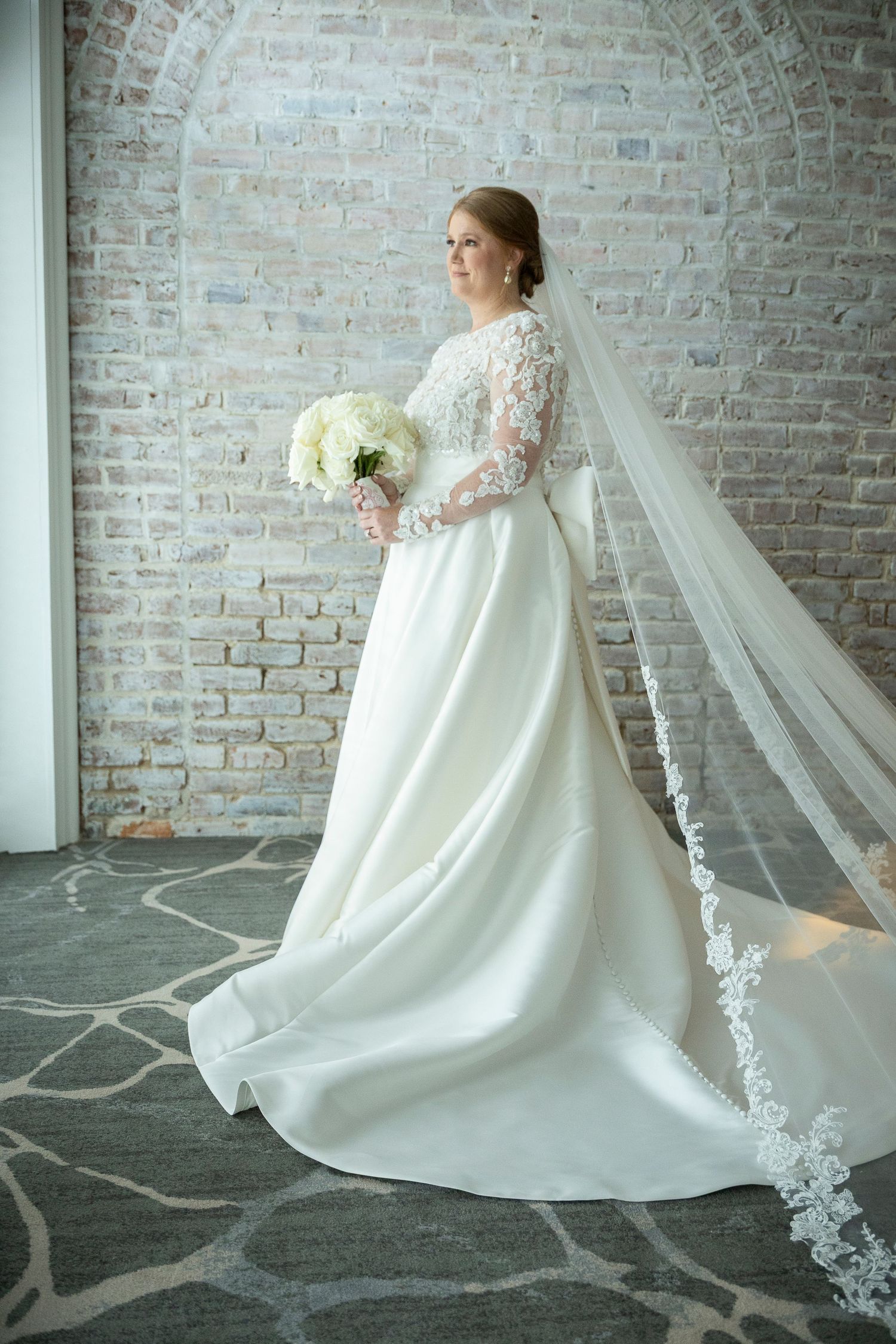 Bride in a white lace long-sleeve ball gown with cathedral veil holds bouquet, posing before rustic brick arch.