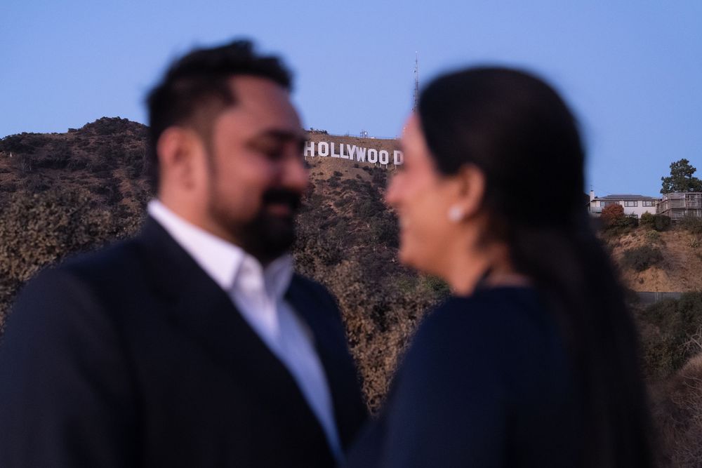 A couple shares a romantic moment with the iconic Hollywood sign visible in the blurred background.